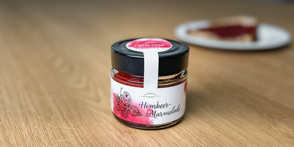 Ein Glas mit Himbeermarmelade und im Hintergrund eine Scheibe Brot mit Marmelade bestrichen